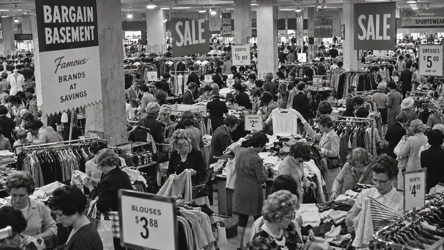 Crowded department store bargain basement with shoppers browsing racks of clothing under signs advertising sales and specific prices.
