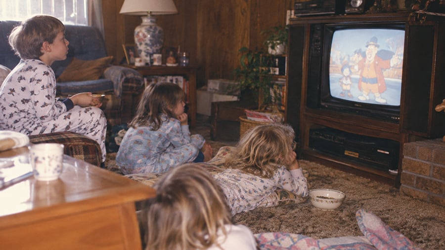 Four children in pajamas watch a cartoon on a box TV in a cozy, wood-paneled living room. They sit or lie on a carpeted floor, surrounded by snacks and cups on a coffee table.