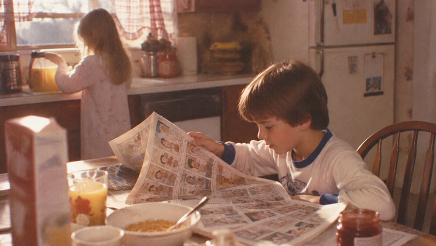 A young boy reads a comic newspaper at a kitchen table with breakfast food, while a girl stands at the sink in the background with sunlight streaming in.