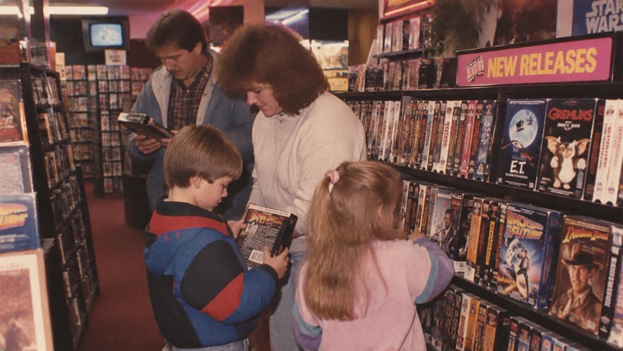 A family browses VHS tapes in a video rental store. Two children and two adults look at movie covers. Shelves are filled with tapes, and a “New Releases” sign is visible above the movie section.