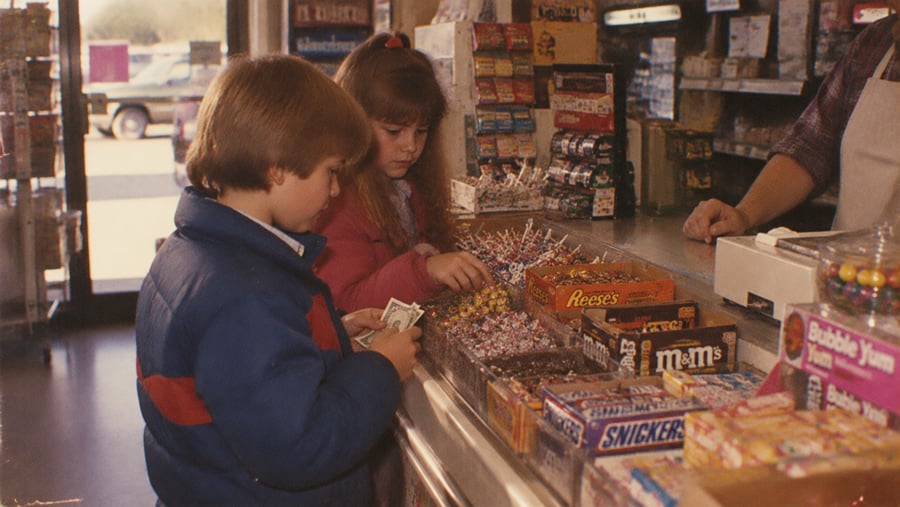 Two children at a store counter choose candy while one holds cash. An assortment of candy bars and lollipops is displayed, and an adult stands behind the counter. Sunlight is visible through the door.