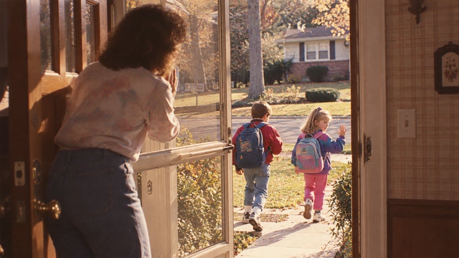 A woman stands at an open door, watching as two young children with backpacks walk down the front path toward school on a sunny day.