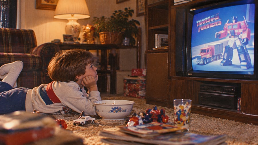 A young child lies on the carpet watching “The Transformers” cartoon on TV. Toys, a bowl, and a drink are nearby, and the room has cozy, vintage decor with warm lighting.
