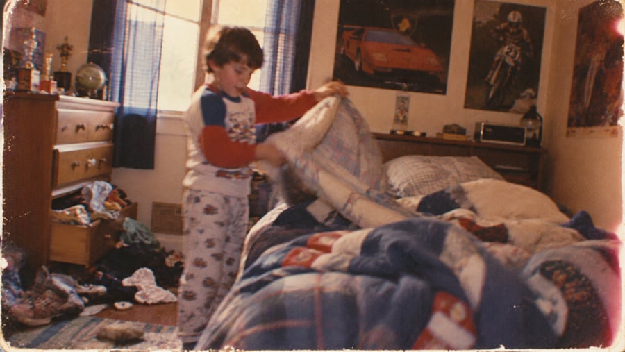 A child in pajamas makes a bed in a messy bedroom with posters on the wall, blue curtains, and scattered clothes on the floor.