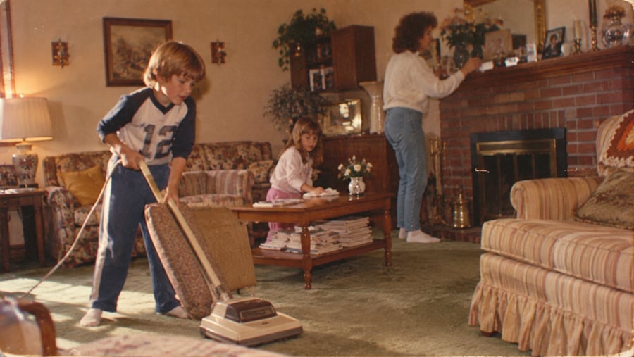 A young boy vacuums a living room while a girl sits on a table and a woman dusts the mantle. The room is decorated with flowers, framed pictures, and cozy furniture.