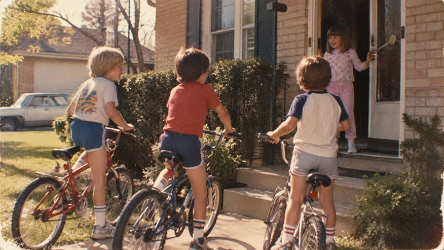 Three children on bicycles face a girl standing at the front door of a brick house on a sunny day. A parked car and greenery are visible in the background.