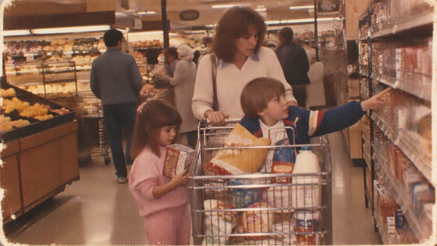 A woman shops with two children in a grocery store. The boy sits in the cart, reaching for an item, while the girl stands nearby holding a box. Shelves and produce are visible in the background.