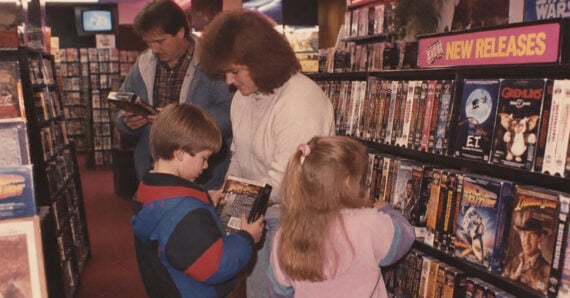 A family browses VHS tapes in a video rental store. Two children look at movies, while a woman and a man check shelves nearby. The “New Releases” sign is visible above a display.