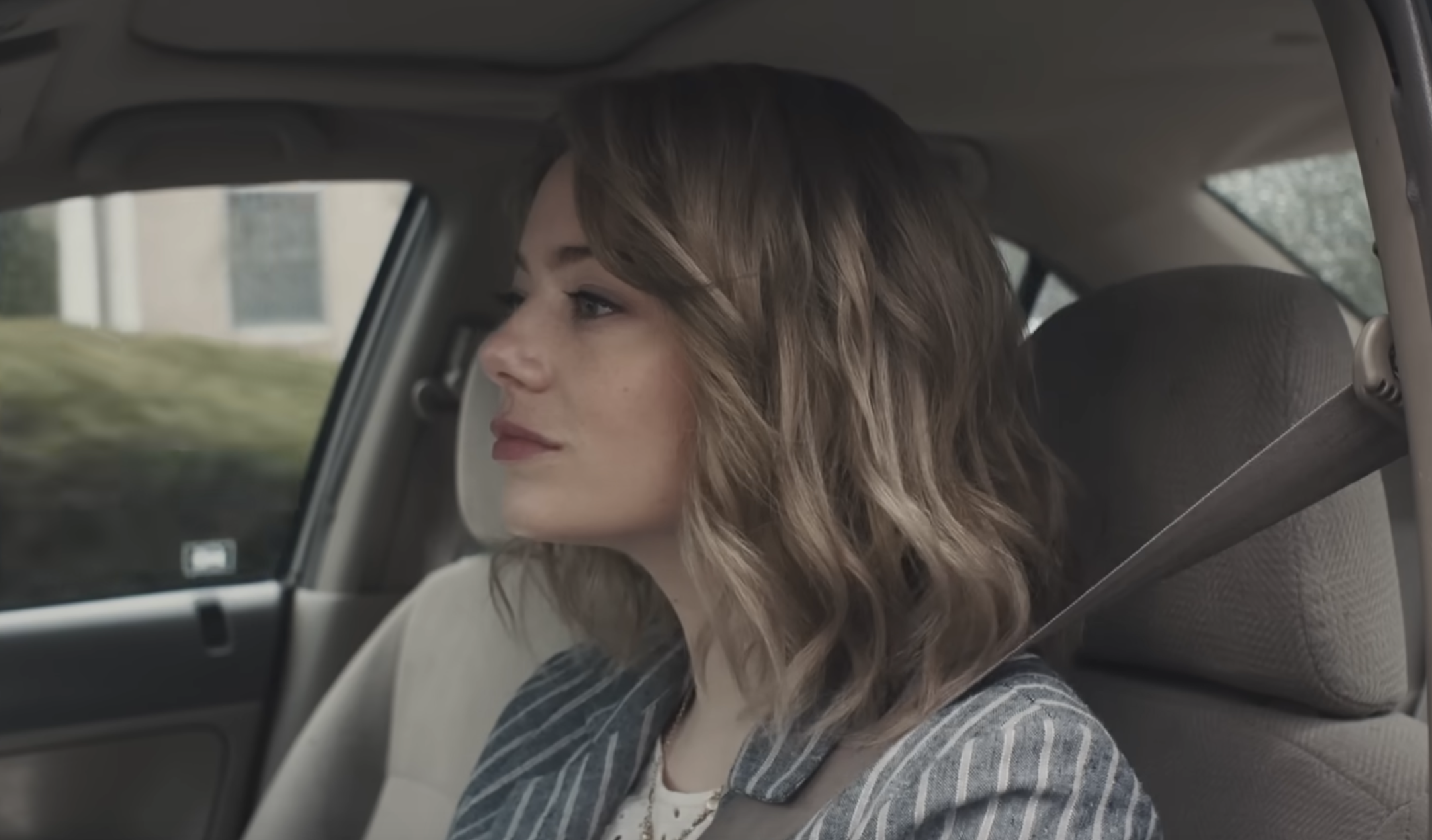 A woman with wavy hair wearing a striped blazer sits in the passenger seat of a car, looking out the window.