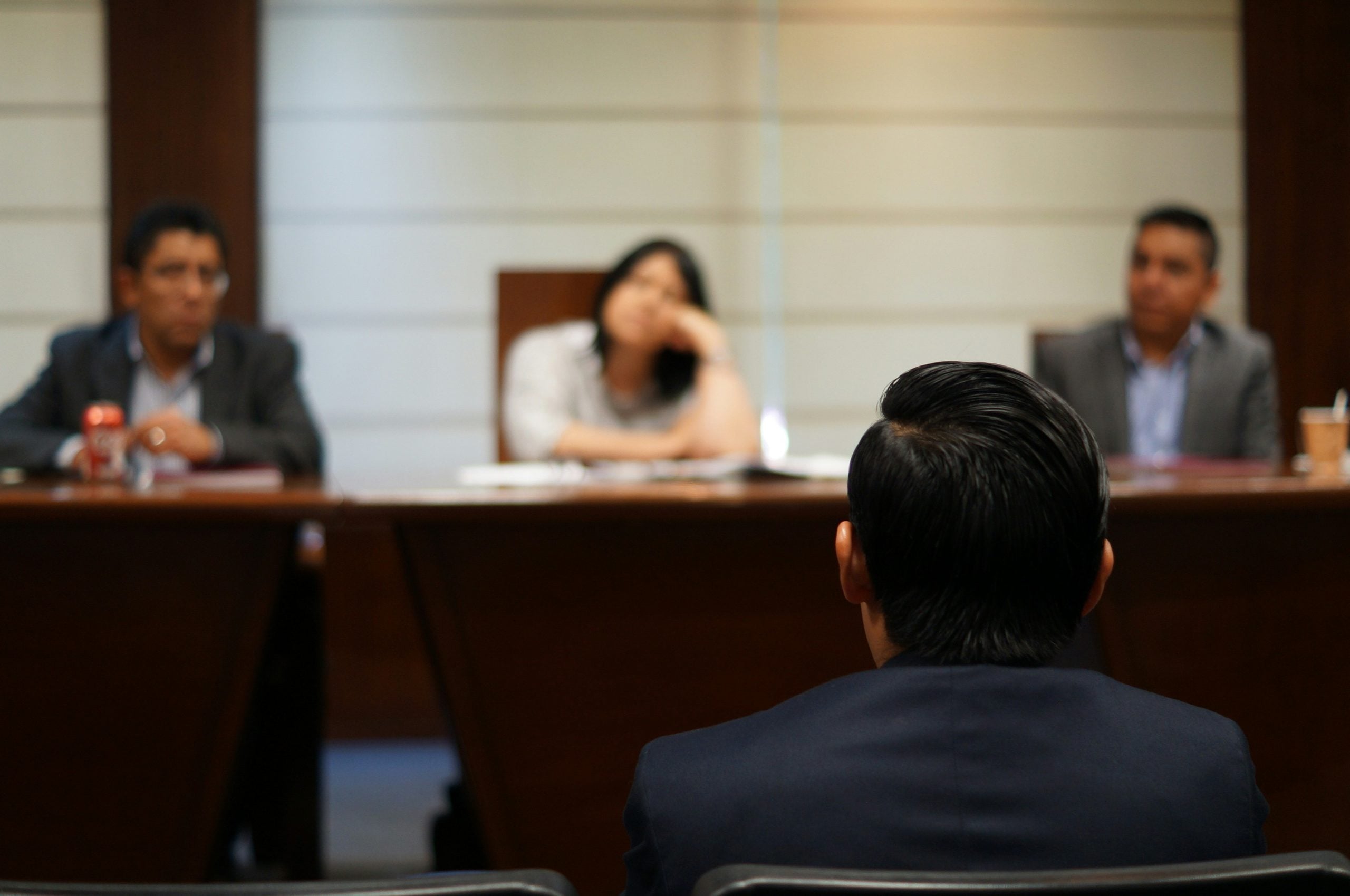 A person in a suit sits in the foreground facing three people seated at a long table during a meeting.