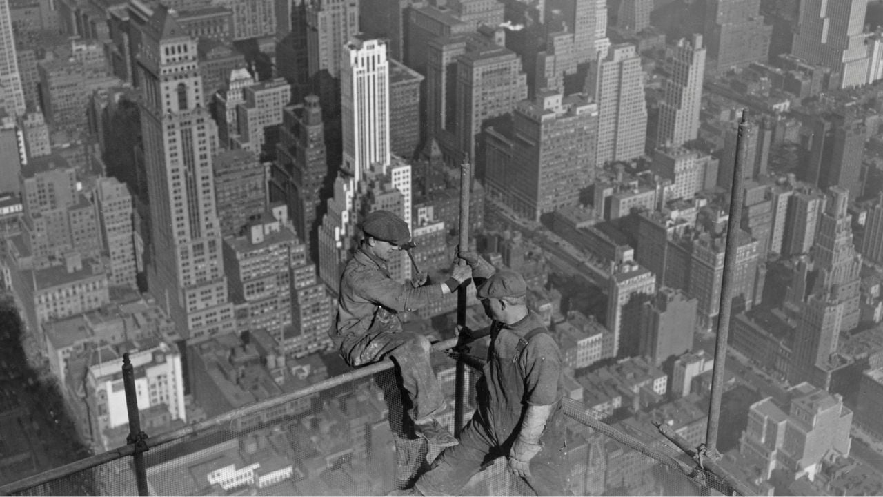 Two construction workers sit on a steel beam high above a city, with skyscrapers and buildings visible below them.