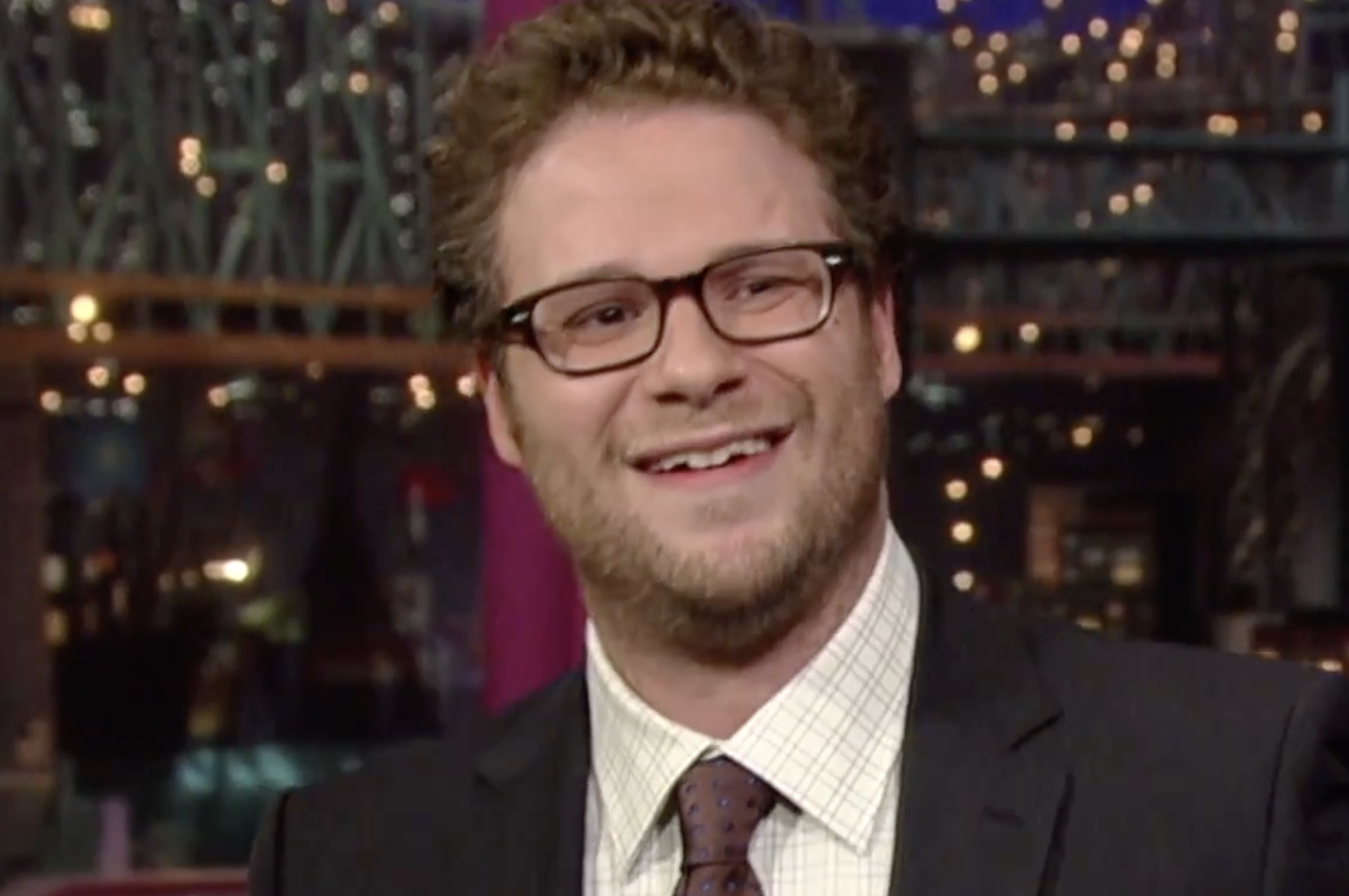 Seth Rogen smiling while wearing a suit, dress shirt, and glasses.