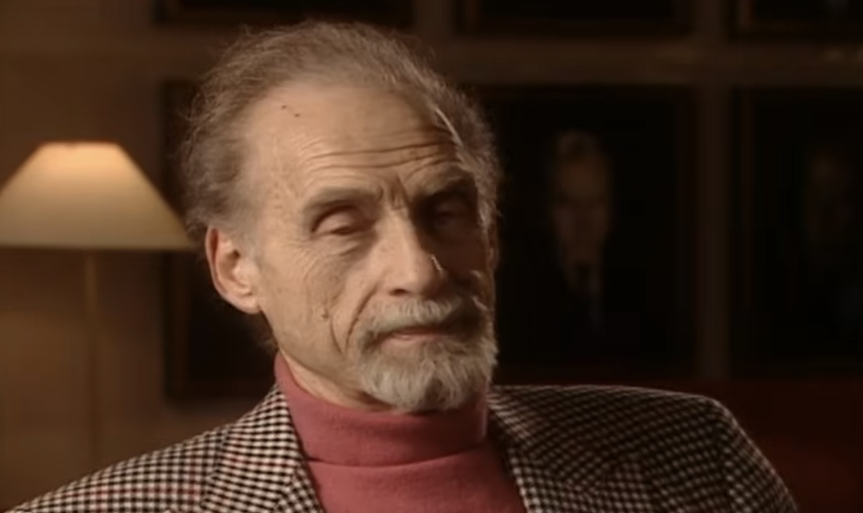 An older man with a gray goatee, wearing a pink turtleneck and a black and white houndstooth blazer.