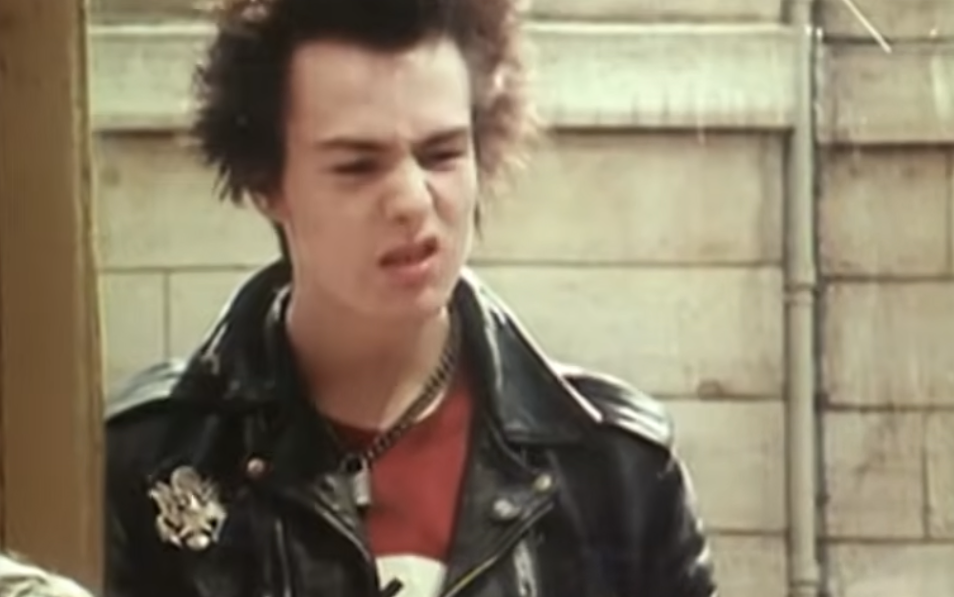 Sid Vicious with spiky hair, wearing a black leather jacket over a red t-shirt, standing outdoors.