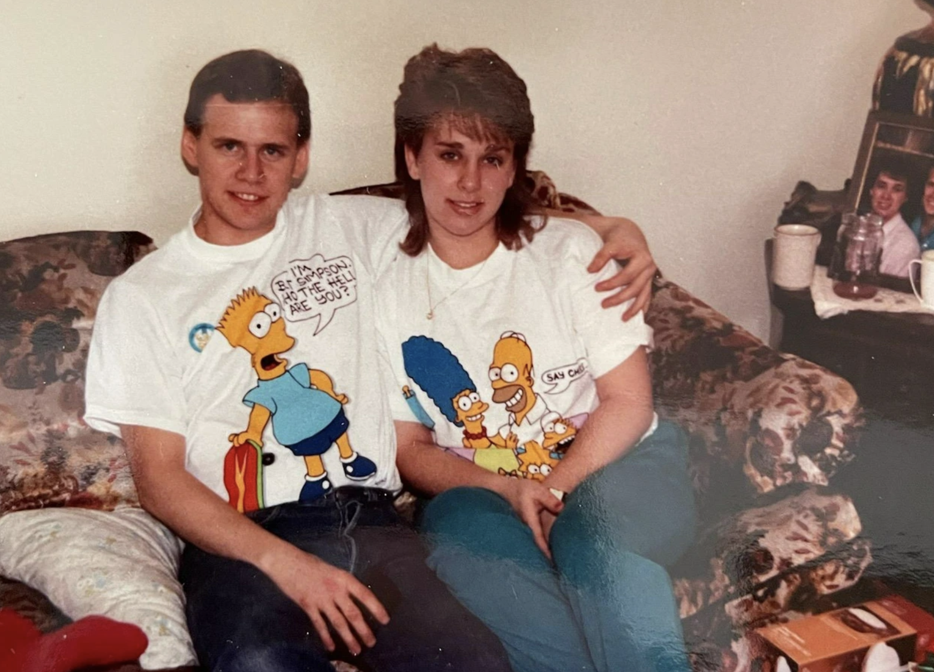 A young man and woman sitting on a couch, both wearing white t-shirts featuring characters from The Simpsons.