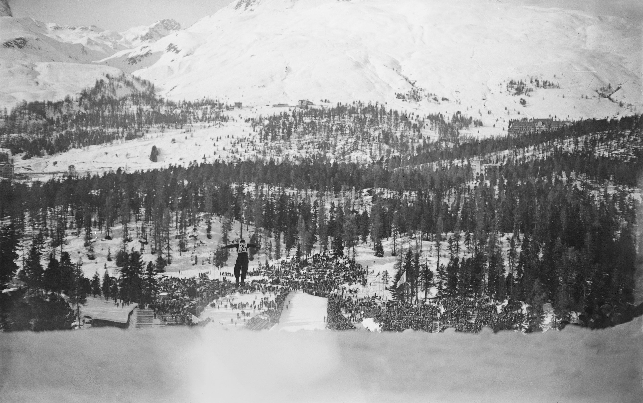 A ski jumper soars above a snowy landscape with a crowd gathered below, surrounded by snow-covered trees and mountains in the background.