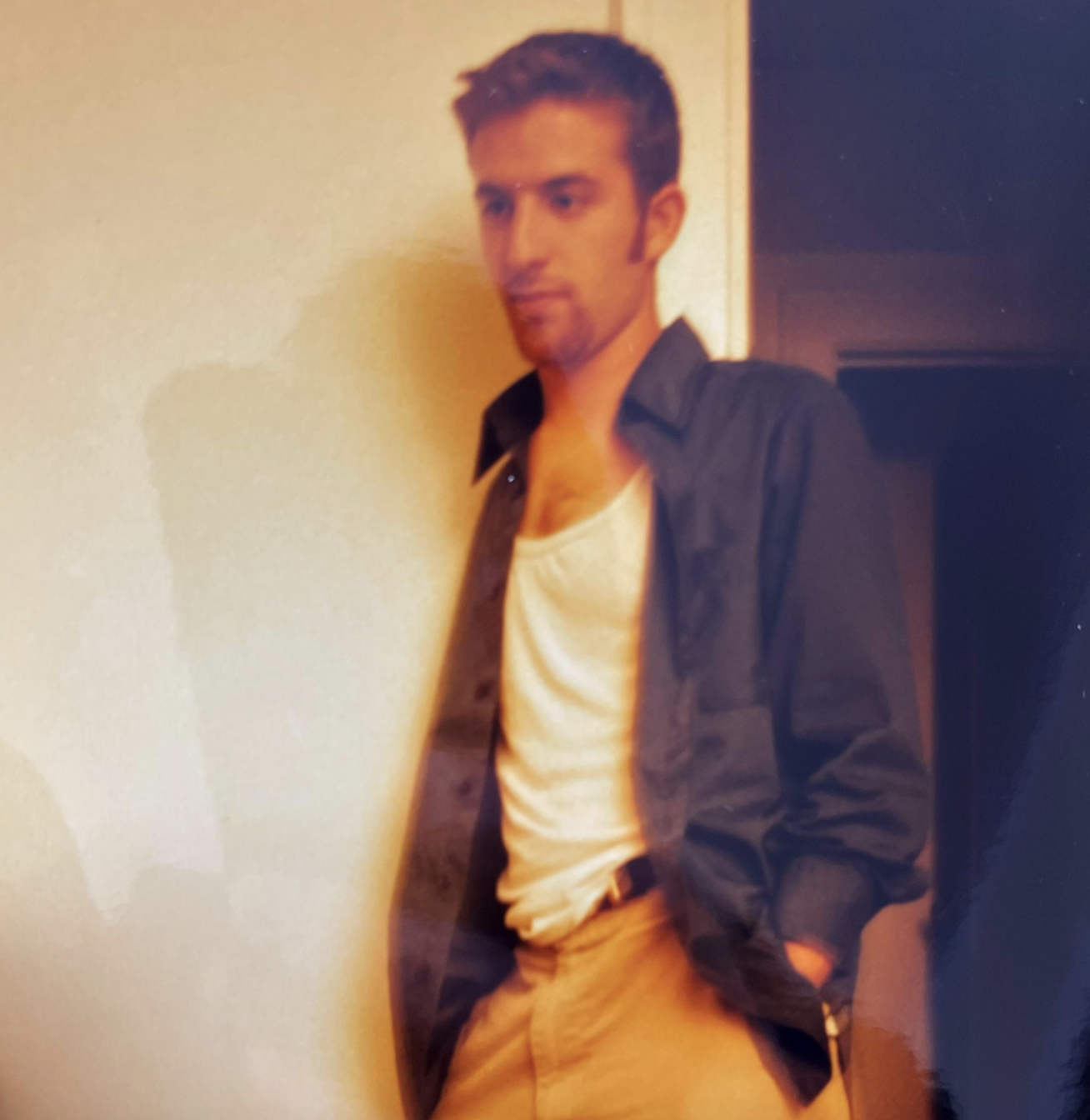 A young man with short dark hair stands wearing an unbuttoned dark shirt over a white tank top and tan trousers.