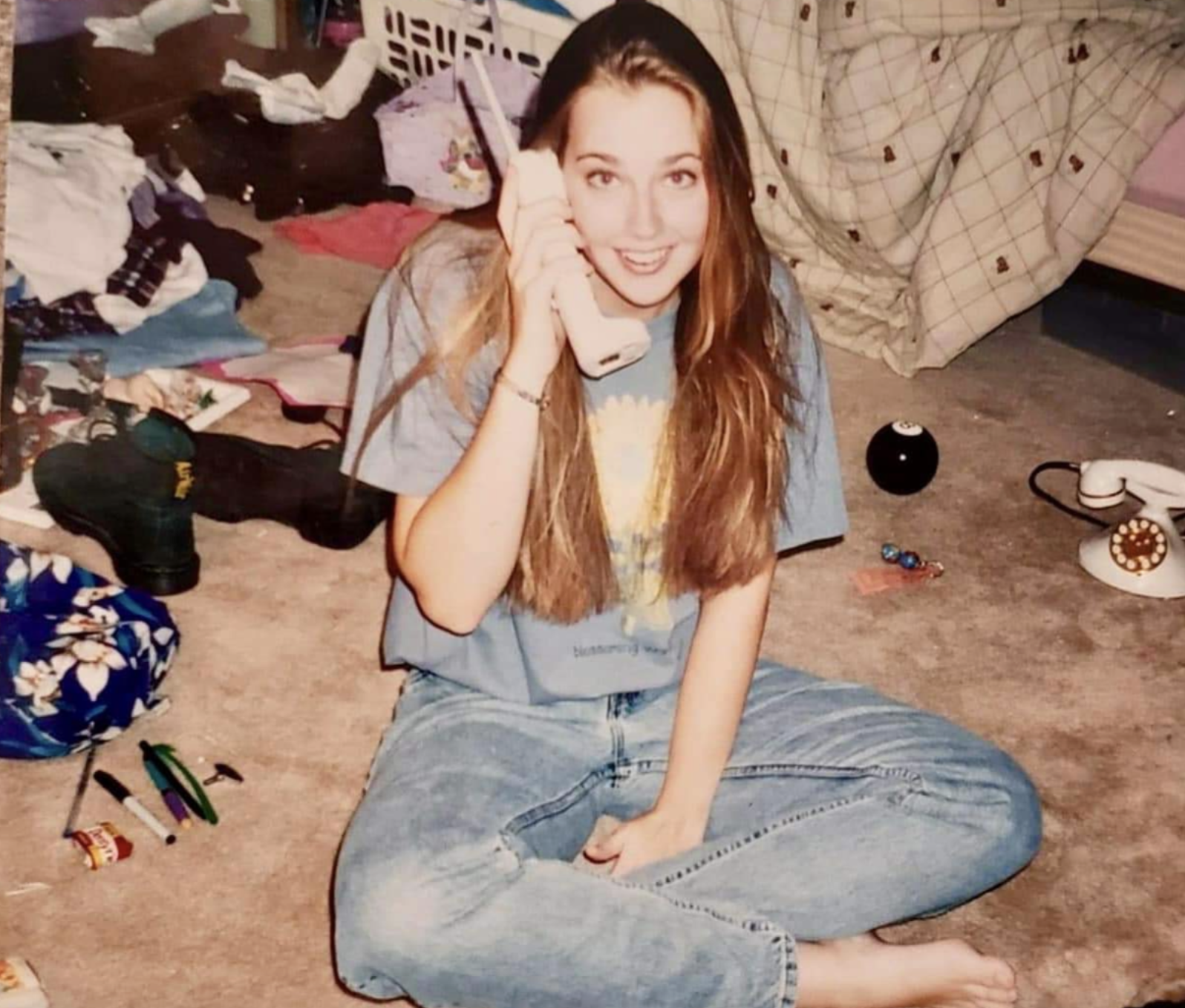 A young woman with long hair sitting on the floor, holding a telephone receiver to her ear and smiling.
