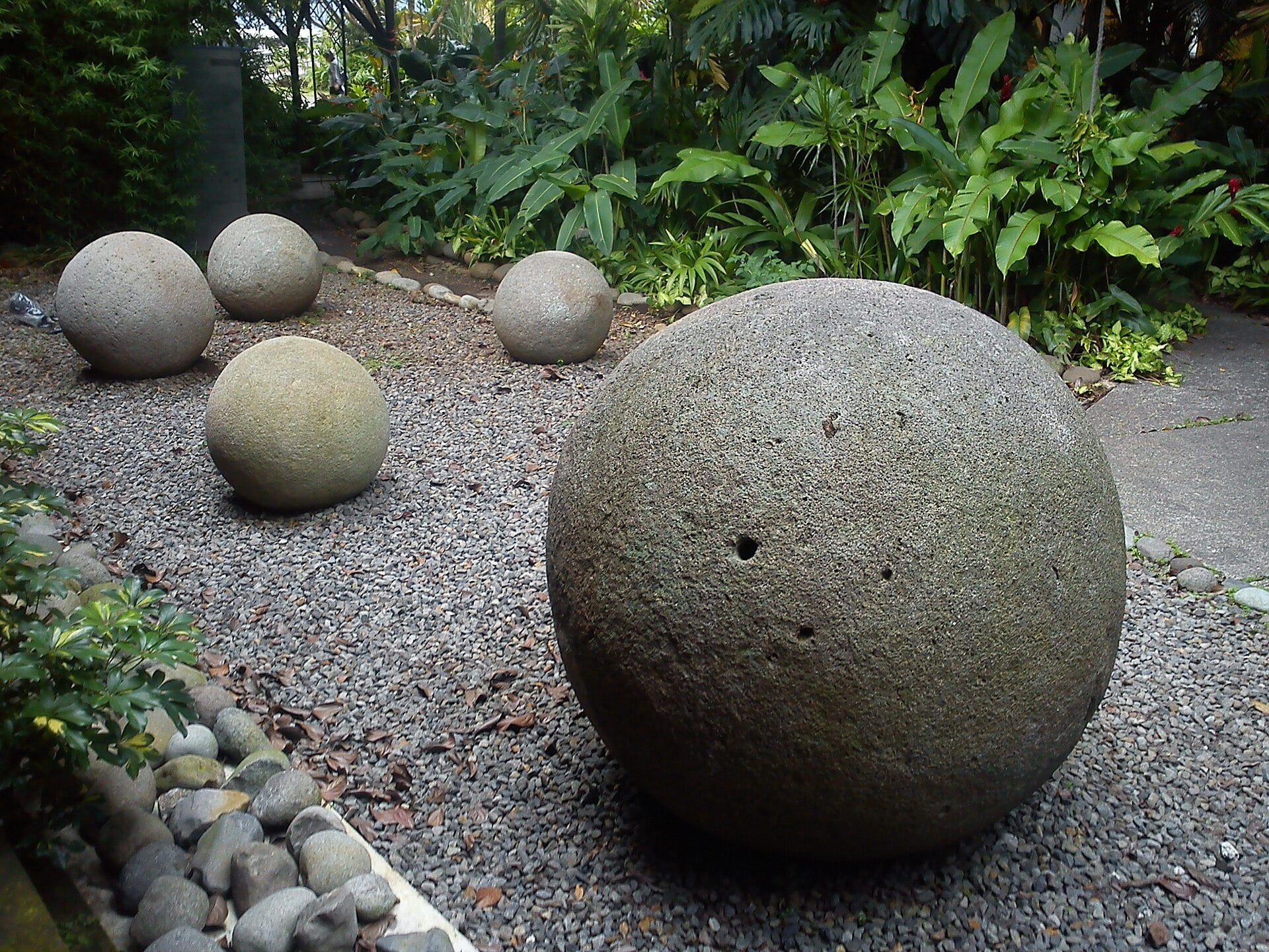 Several large, spherical stone sculptures resting on a gravel garden path surrounded by tropical plants.
