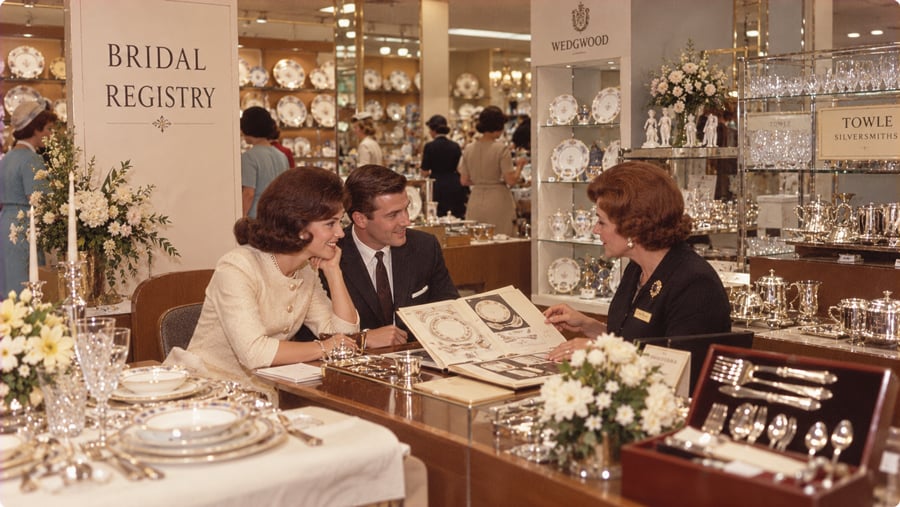 A couple at a bridal registry counter looks at a catalog with a sales associate, surrounded by displays of fine china and silverware.