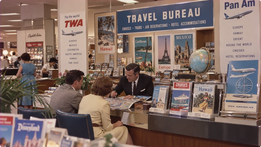 A travel agent sits at a desk with a couple, surrounded by vintage posters and displays for TWA and Pan Am.