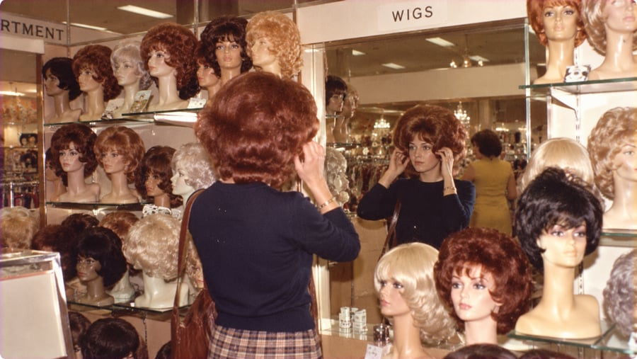 A woman with a bouffant hairstyle stands in front of a mirror in a store aisle lined with shelves of mannequin heads wearing various wigs.