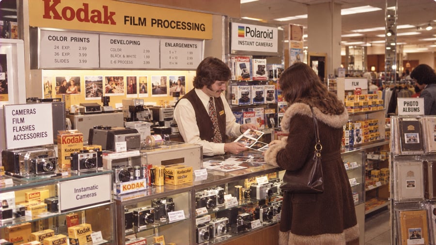 A clerk in a vest shows developed photographs to a customer at a store counter filled with Kodak cameras and film processing price signage.