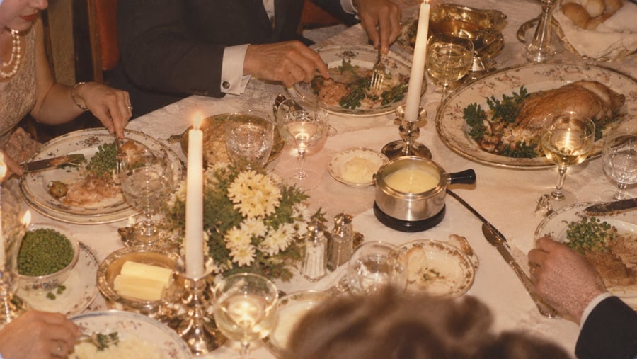 A group of people dining at a formal table set with a roasted chicken, peas, fondue, butter, and lit candles.