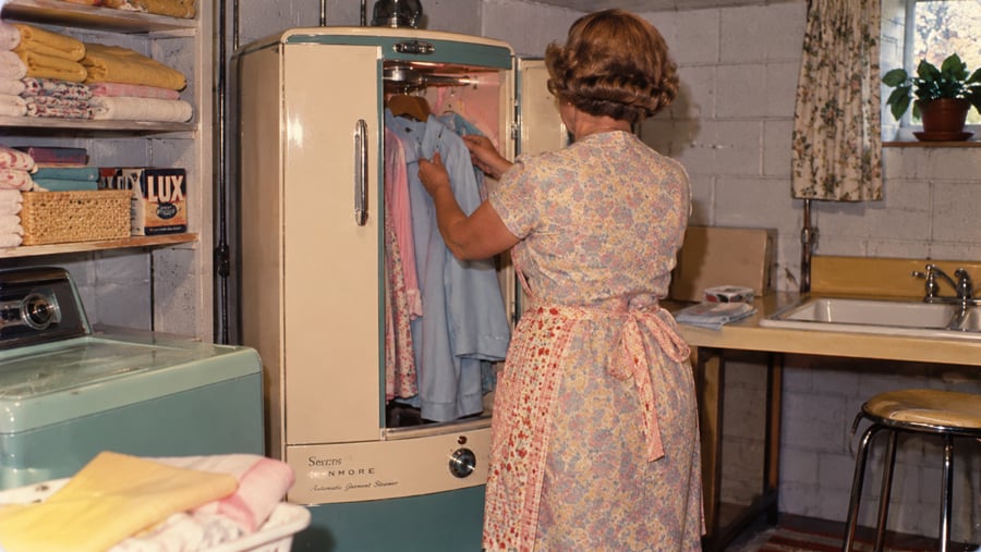 A woman in a floral dress stands in a laundry room, placing a garment into a Kenmore automatic garment steamer.