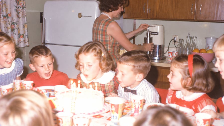 A young girl blows out candles on a birthday cake at a table surrounded by children, while a woman prepares drinks in the background.