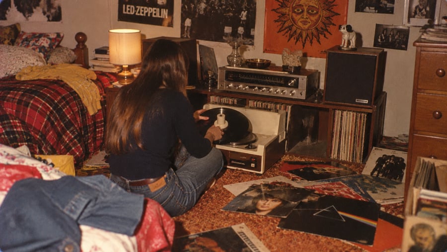 A person sitting on a carpeted floor in a bedroom, placing a vinyl record onto a turntable surrounded by music albums and posters.