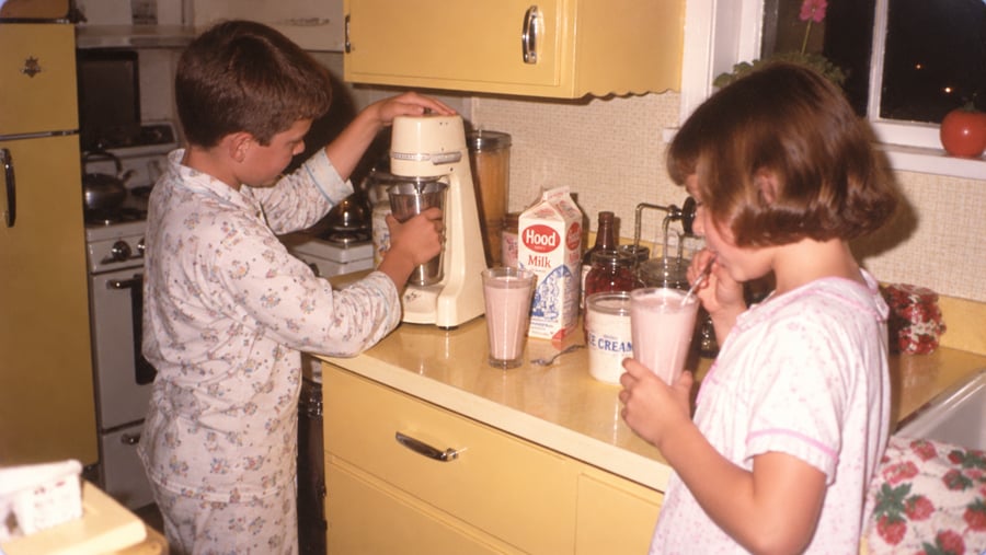 A boy in pajamas uses an electric stand mixer to make a milkshake while a girl in pajamas drinks a glass of milk on a kitchen counter.