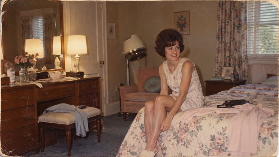 A smiling woman with dark hair sits on the edge of a floral bedspread in a bedroom, with a vanity and a floor-standing hair dryer in the background.