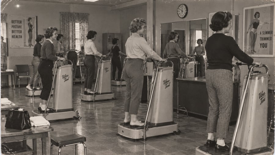 Several women stand on vibrating belt machines in a gym, with health-focused posters on the walls.