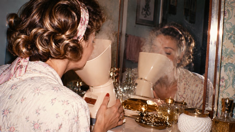 A woman in a floral robe uses a facial steamer in front of a mirror.
