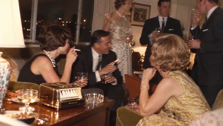 A group of people in formal attire socializing, drinking, and smoking in a dimly lit room.