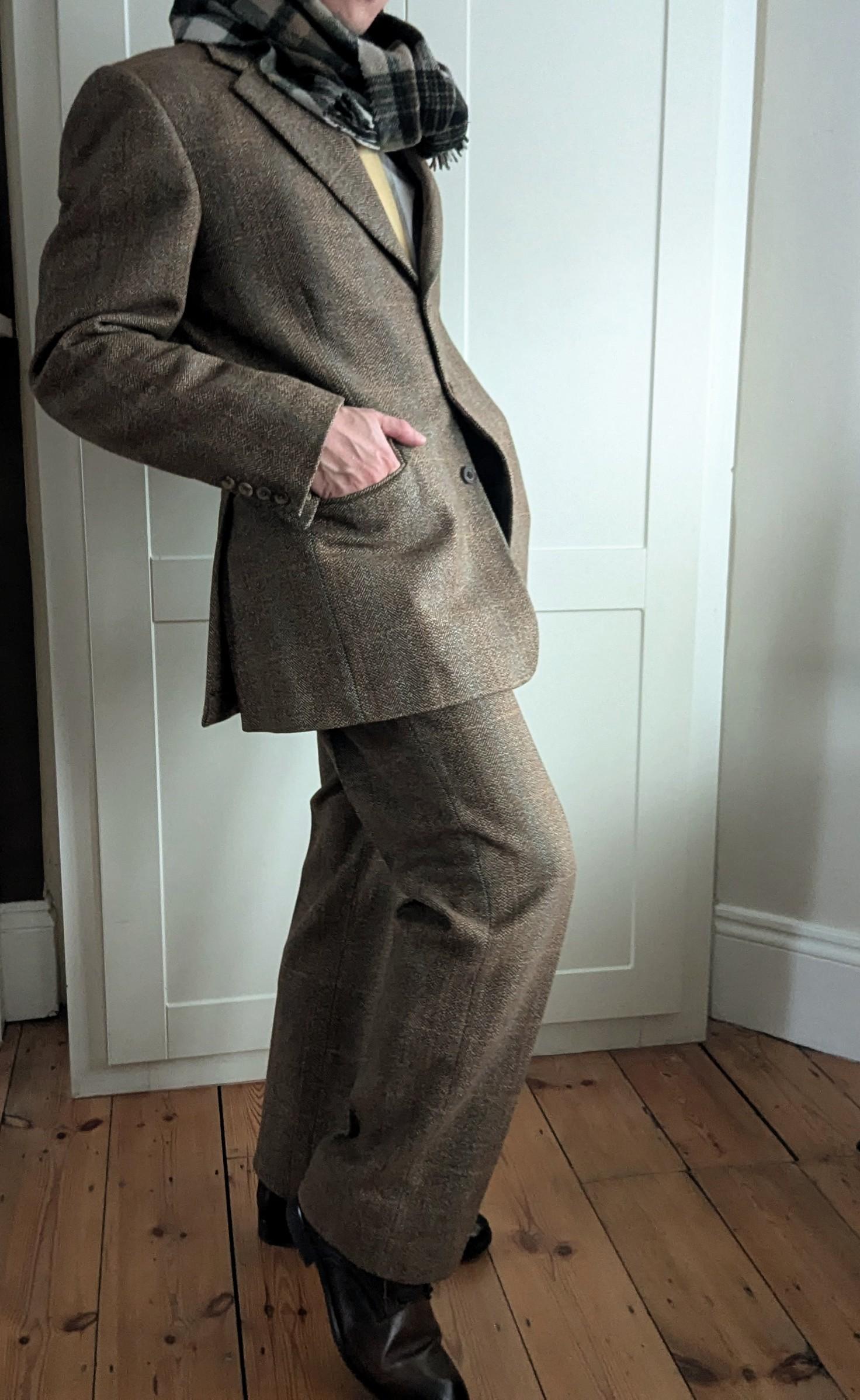 A person poses indoors wearing a brown plaid suit, black shoes, and a black-and-gray checkered scarf, with one hand in their pocket and the other arm bent.