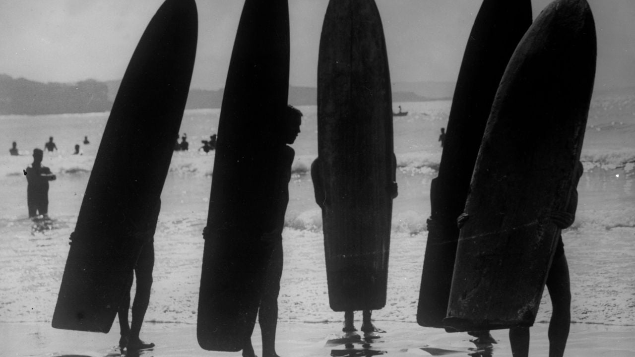 Five people stand on a beach holding large surfboards upright, silhouetted against the ocean. Other beachgoers are visible in the background, and the scene appears to be set on a sunny day.