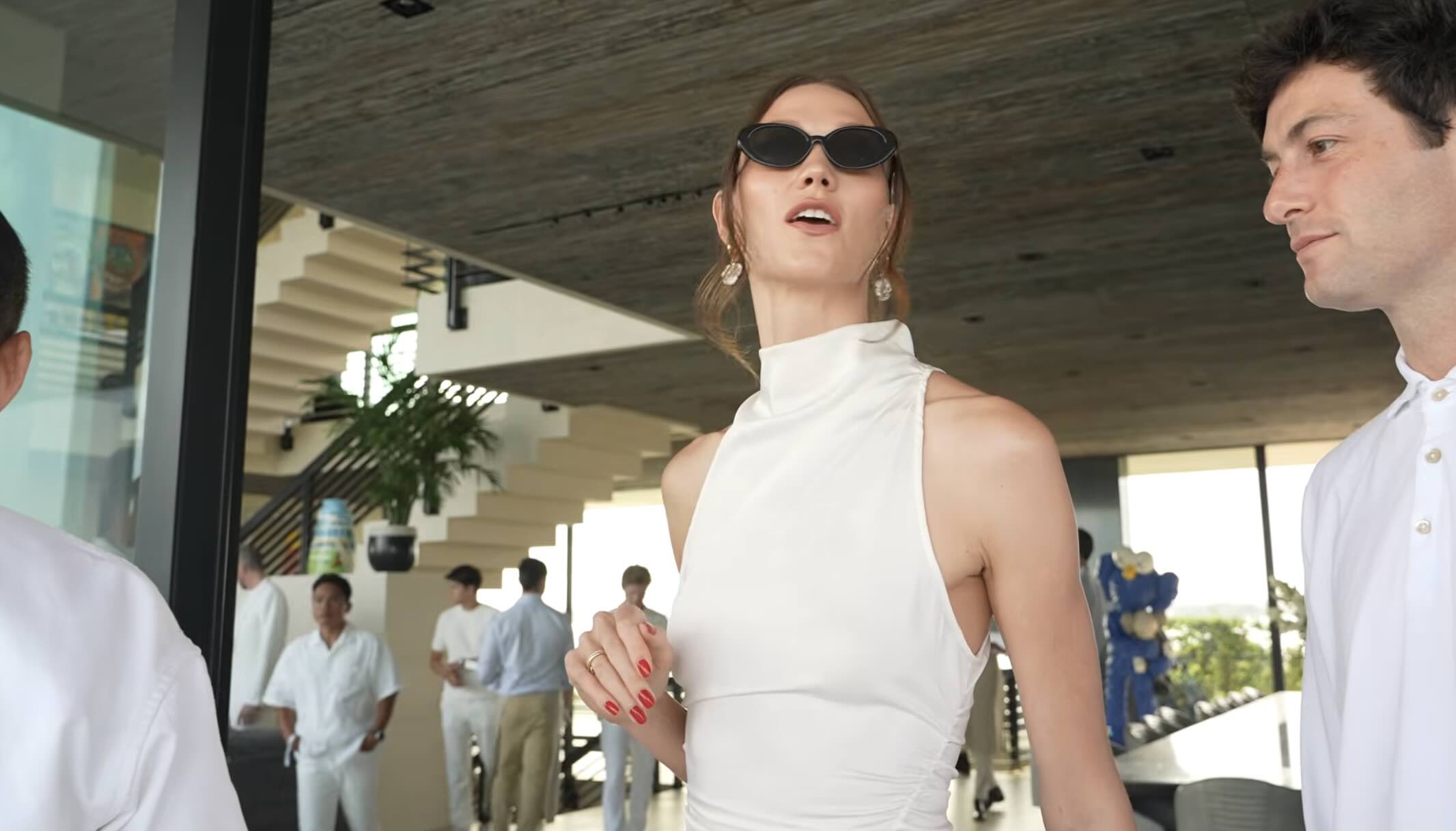 A woman wearing sunglasses and a white sleeveless top stands in a bright, open-plan space with a man, while others socialize in the background.