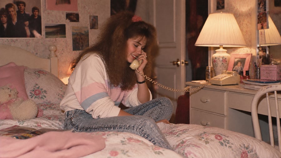 A young woman with voluminous hair sits on a floral bed, smiling while talking on a corded telephone in her bedroom.