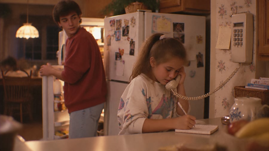 A girl at a kitchen counter talks on a wall-mounted telephone while writing in a notebook, as a young man stands by the open refrigerator behind her.