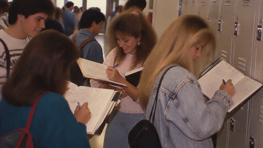 Four students stand in a school hallway, writing in notebooks held against a row of lockers.
