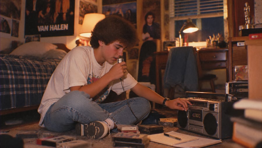 A teenager sits on the floor of a bedroom, holding a microphone while adjusting a cassette boombox surrounded by cassette tapes.