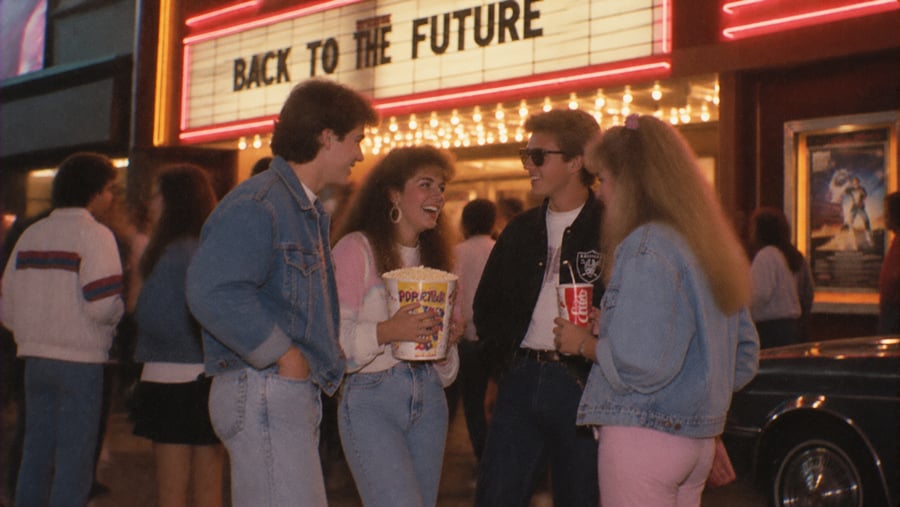 Four young adults in 1980s attire standing in front of a movie theater marquee advertising “Back to the Future.”