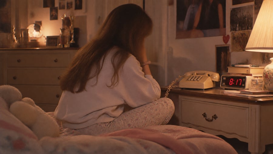 A person sits on a bed with their head in their hands next to a bedside table with a rotary phone and a digital clock displaying 8:17.