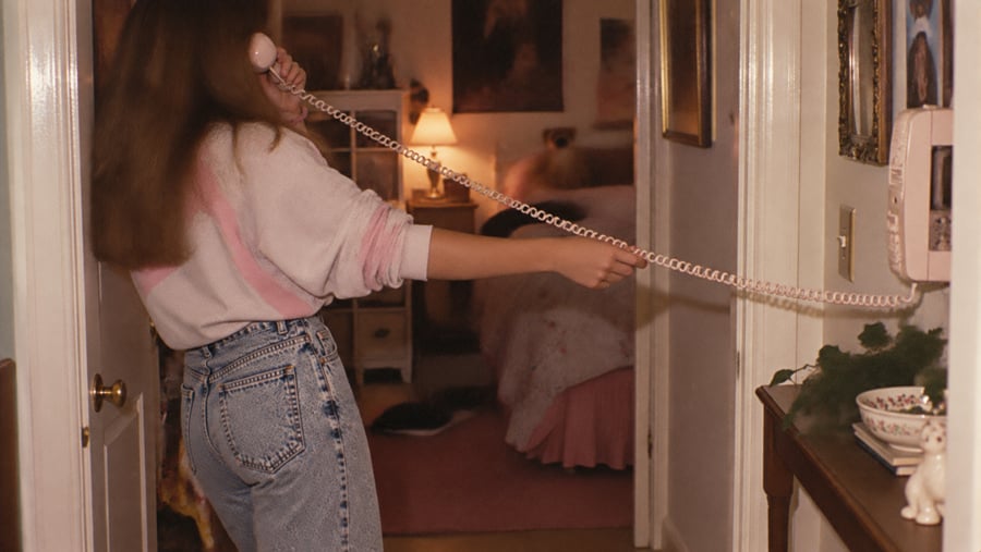 A person with long hair stands in a doorway, holding a wall-mounted telephone handset to their ear while stretching the long, coiled cord across the hallway.