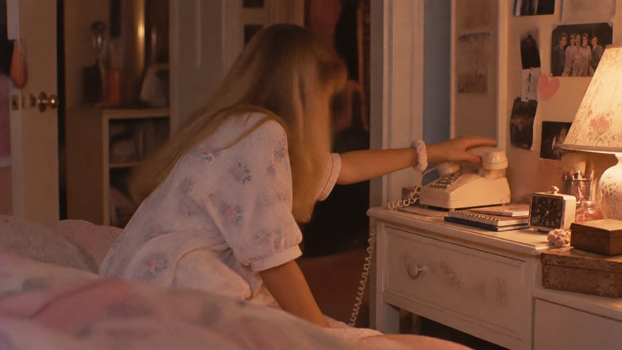 A young woman with long blonde hair sits on her bed and reaches toward a vintage rotary telephone on a bedside table.