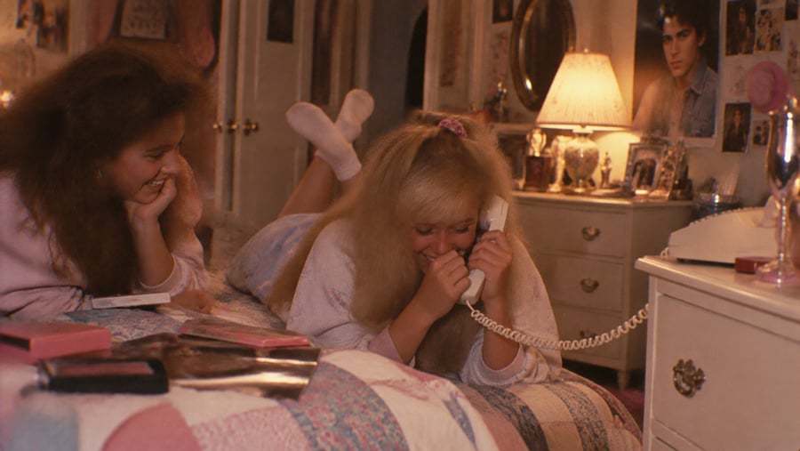 Two teenage girls in pajamas laugh while one talks on a corded landline phone while lying on a bed.