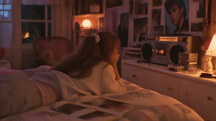 A girl lying on a bed listens to a portable cassette player in a dimly lit room.