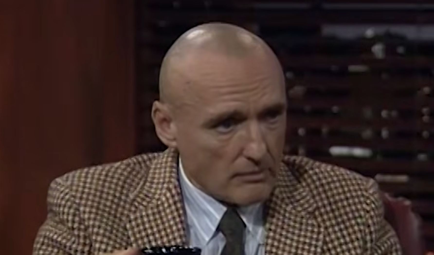 A bald man with a serious expression wears a brown plaid blazer, a striped button-down shirt, and a dark tie.